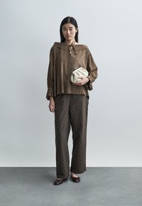 Καφέ σουέτ τοπ με γιακά και λεπτομέρεια κορδονιού, συνδυασμένο με καφέ παντελόνι καμπάνα με σχέδια και κρεμ τσάντα χειρός. Καφέ φλατ παπούτσια ολοκληρώνουν το σύνολο.