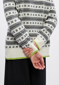 Strikkegenser med svart og hvitt geometrisk mønster, ribbestrikkede mansjetter og grønn aksent langs kanten og ermekantene.