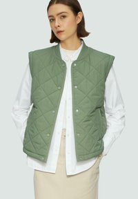 Gilet matelassé vert avec des boutons-pression, doté d'un col rond et sans manches, superposé à une chemise blanche à boutons. Texture de tissu uni.