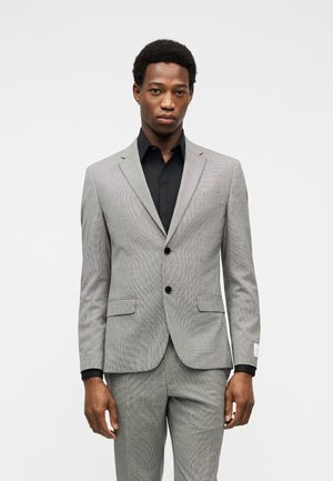 Young man wearing a light gray suit with black buttons and a black dress shirt, standing against a plain white background.