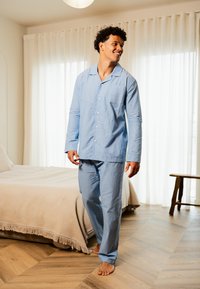 Ensemble de pyjama rayé bleu clair avec manches longues et pantalon, comportant une poche sur le haut. Intérieur de la pièce avec un sol en bois et un lit.