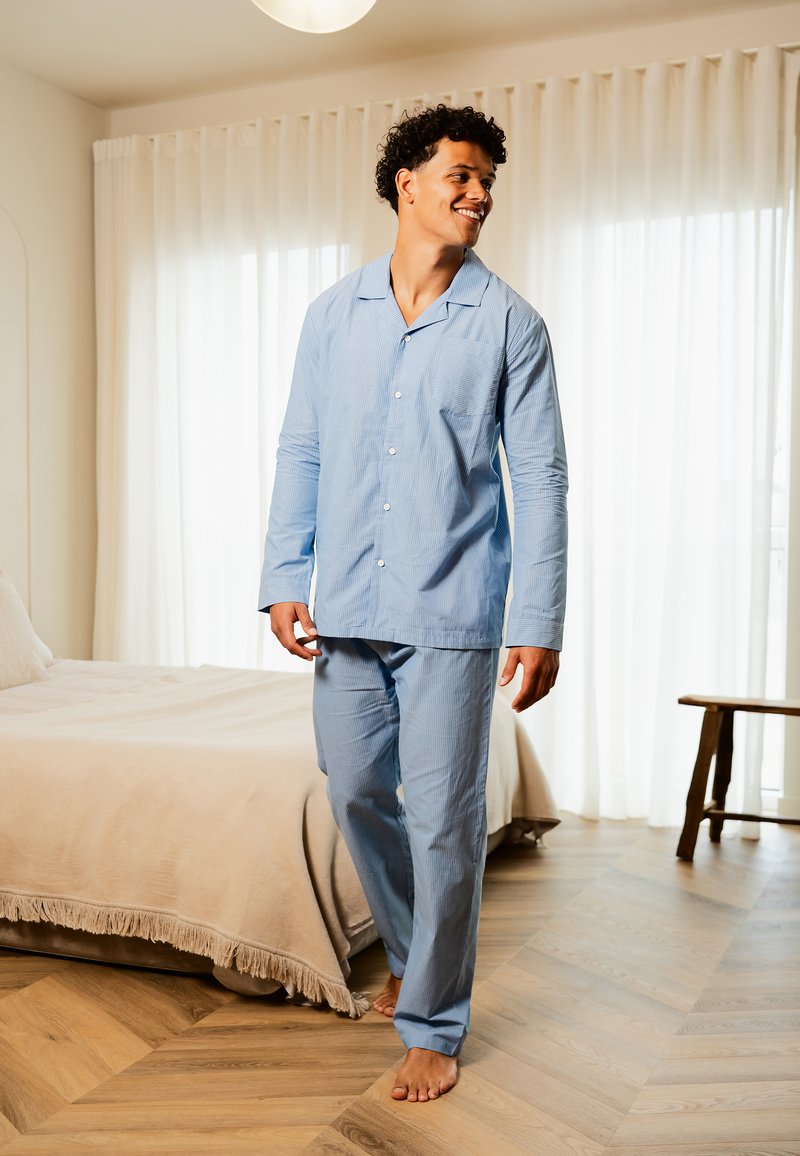 Ensemble de pyjama rayé bleu clair avec manches longues et pantalon, comportant une poche sur le haut. Intérieur de la pièce avec un sol en bois et un lit.