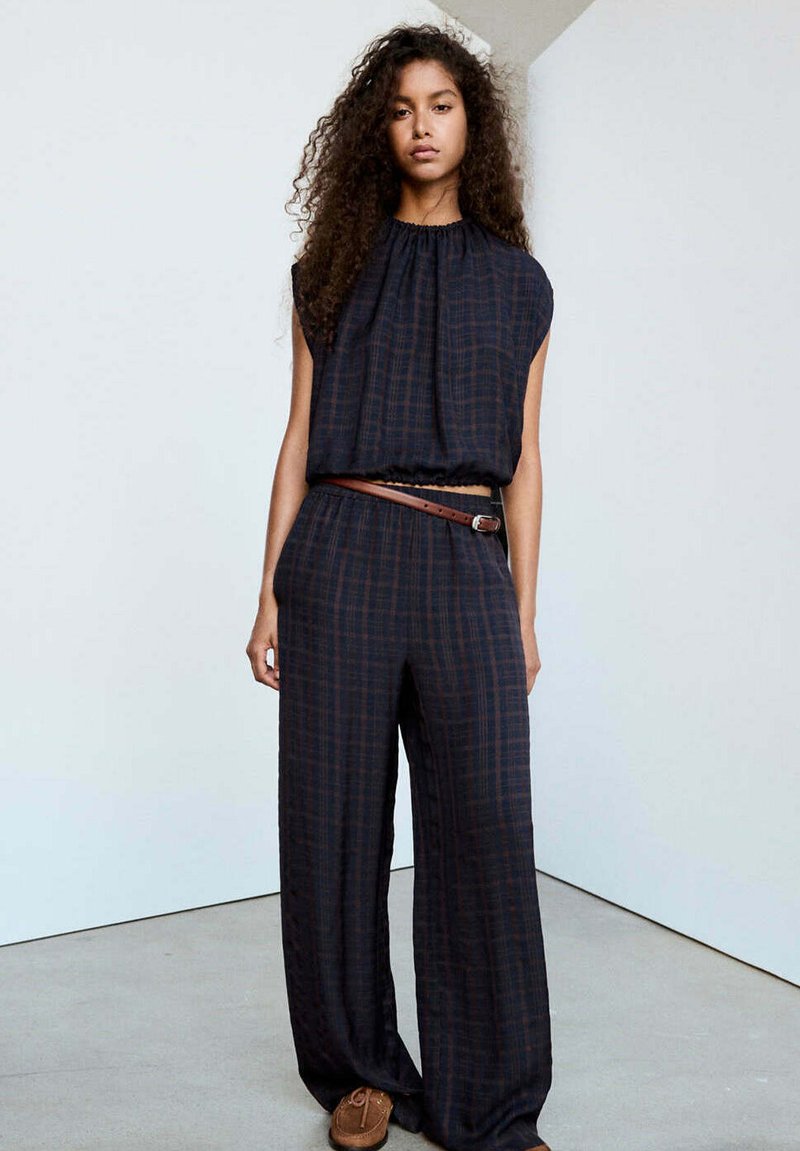 Femme aux cheveux bouclés portant un haut sans manches ample à carreaux foncés et un pantalon large, avec une ceinture marron et des mocassins marrons.