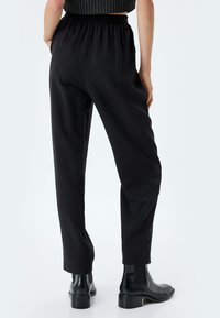 Pantalons noirs à taille élastique avec une coupe droite. Le matériau a une texture lisse. Portés avec des bottines noires élégantes.