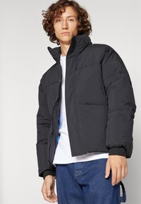 Schwarze Puffjacke mit hohem Kragen, Reißverschluss und zwei Fronttaschen. Der Stoff wirkt glatt mit einer leicht strukturierten Oberflächenbeschaffenheit.
