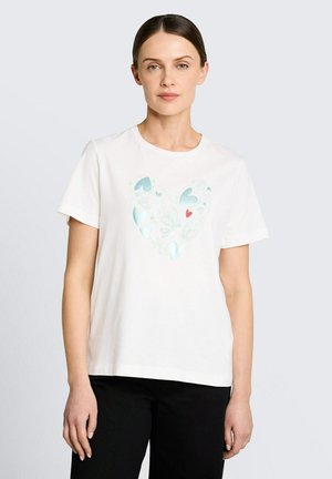 Kobieta o ciemnych włosach w białym t-shircie z dużą grafiką serca w niebieskich i czerwonych elementach, ubrana w czarne spodnie, na jednolitym tle.