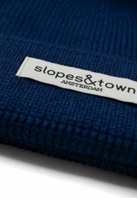 Beanie lavorato a maglia blu navy con texture a coste, dotato di un'etichetta bianca con la scritta "slopes&town AMSTERDAM". Il design è semplice e pratico.