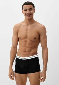 Smiling muscular man wearing black boxer briefs with white waistband featuring "QS" logo, standing against plain light background.