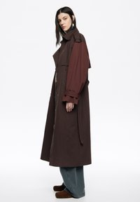 Długi, dwurzędowy płaszcz z blokowym wzorem w ciemnobrązowym i bordo kolorze, z paskiem w talii i szerokimi rękawami. Noszony na luźnych dżinsach.
