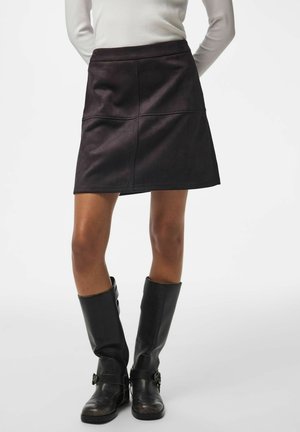 Femme portant une mini-jupe en cuir noire, un haut blanc à manches longues et des bottes noires montant jusqu'aux genoux, debout les mains derrière le dos.