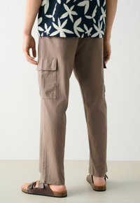 Beige Cargo-Hosen mit Seitentaschen, aus leichtem Material, gerader Schnitt und kombiniert mit braunen Slipper-Sandalen. Der Hintergrund ist neutral.