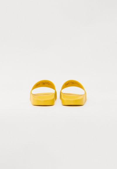 Κίτρινα λάστιχα παπούτσια ολίσθησης με λεία υφή, ευρύ σχέδιο ανοιχτής στεφάνης και παχύ σόλα, σε θέα από το πίσω μέρος σε λευκό φόντο.