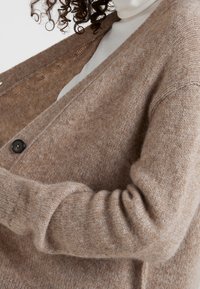 Bruine wollen vest met een losse pasvorm, V-halsontwerp en een enkele zwarte knoopsluiting. Zachte textuur met geribbelde manchetten en zoom.