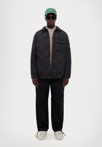 Giacca nera trapuntata con tasca a toppa, abbinata a un maglione beige testurizzato, pantaloni neri e sneaker bianche. Cappello verde e occhiali da sole inclusi.