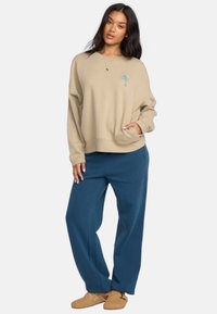 Beżowy sweatshirt z haftem palmy, z przednią kieszenią, zestawiony z niebieskimi spodniami szerokimi i brązowymi butami wsuwanymi.