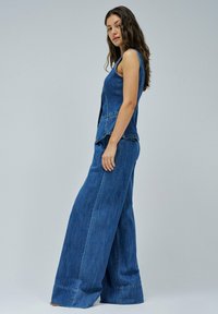 Combinaison en denim bleu foncé, présentant un design à jambes larges, une coupe sans manches et des détails de couture visibles le long des coutures.