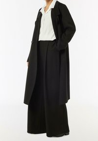 Cappotto nero con un taglio lungo e su misura, dotato di chiusura con un bottone singolo, abbinato a una camicia bianca con colletto e pantaloni neri larghi.