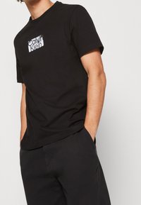 Camiseta de algodón negra con un parche de logo blanco en el pecho, cuello redondo y mangas cortas. El modelo está de pie con las manos en los bolsillos.