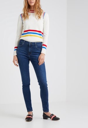 Suéter de base blanca con rayas rojas, azules y amarillas; combinado con jeans ajustados azul oscuro y zapatos negros con acentos rojos y tachuelas.