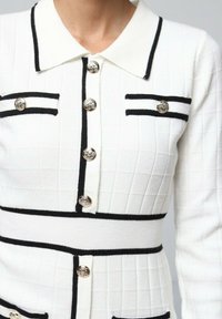 Cardigan en tricot blanc avec un motif en grille, des accents noirs et des boutons argentés. Doté d'un col pointu et de deux poches décoratives.