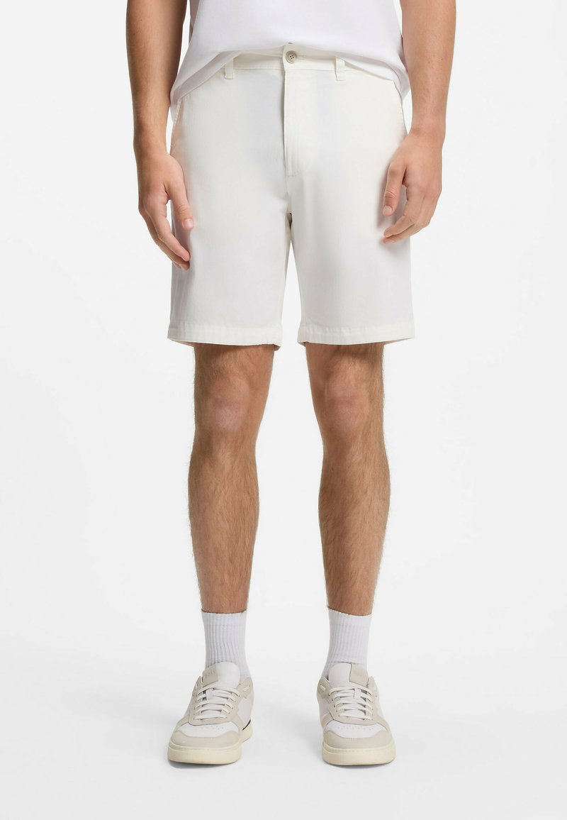 Homme portant un short décontracté blanc, des chaussettes montantes blanches et des baskets blanches, debout les bras détendus le long du corps devant un fond uni.
