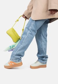 Un jean bleu clair, un blazer beige oversize, des baskets plateformes menthe verte avec des rayures orange, et un petit sac à main jaune avec une poignée nouée.