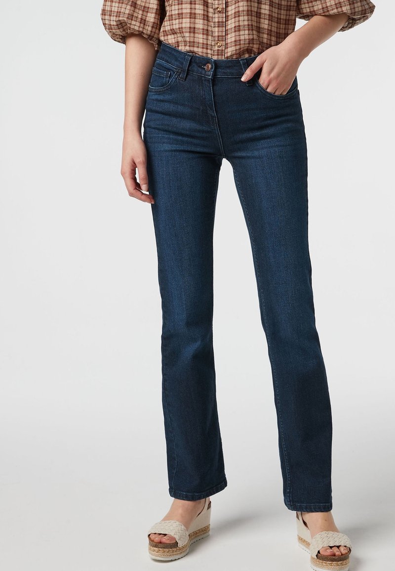 Femme portant un jean droit bleu foncé, une chemise à carreaux et des sandales compensées beiges, debout avec une main dans la poche.