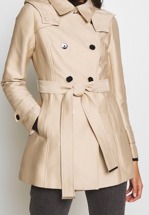 Beige double-breasted trench coat with black buttons and waist belt, worn over dark pants, right hand in pocket.