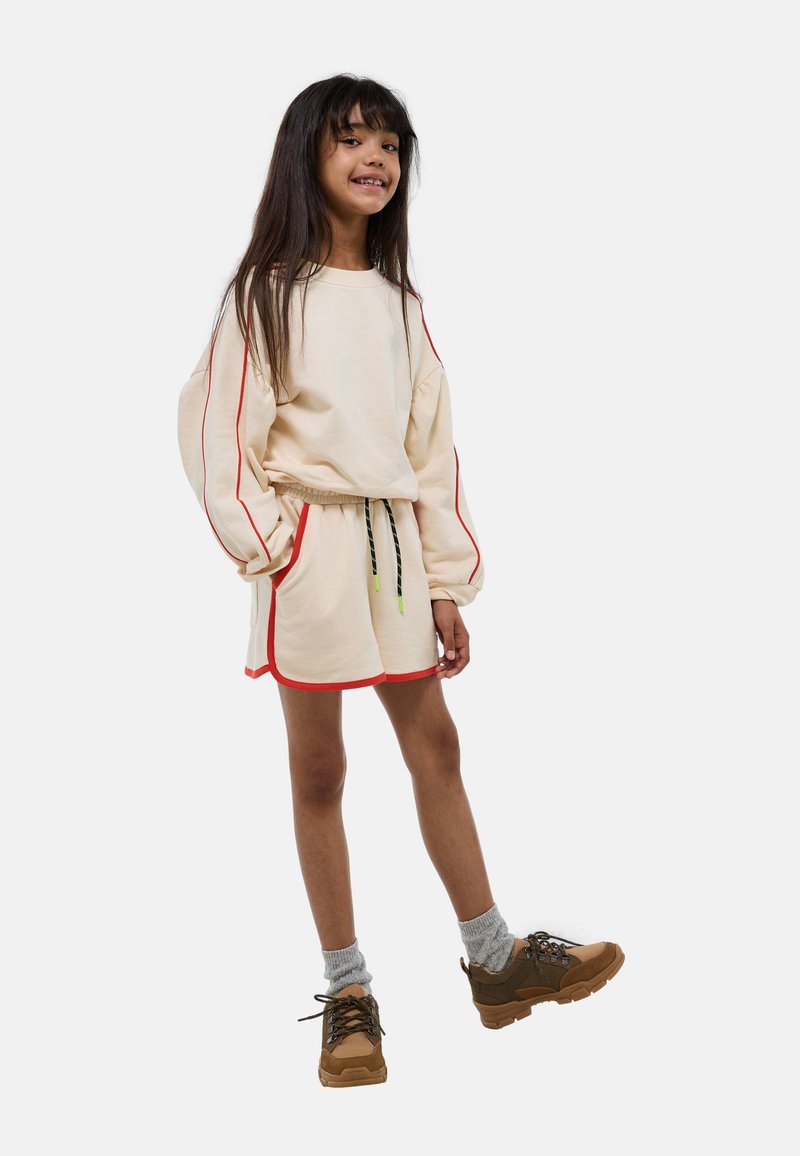 Ensemble en crème composé d'un sweat-shirt et d'un short avec des finitions rouges, doté de manches bouffantes et d'une taille à cordon, associé à des chaussures de randonnée marron et des chaussettes grises.