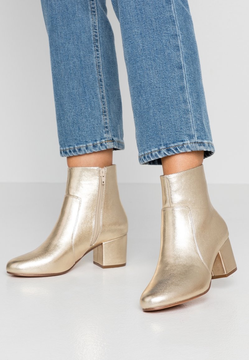 Pieds chaussés de bottines métalliques dorées à talons carrés avec fermetures éclair latérales, associées à un jean bleu court sur un fond uni.