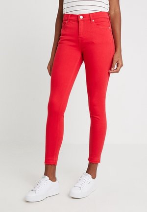 Femme portant un jean skinny rouge vif avec des ourlets bruts, associé à des baskets blanches et un haut blanc rayé.