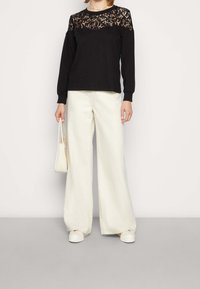 Top nero a maniche lunghe con motivo di pizzo sulle spalle, abbinato a pantaloni bianchi a gamba larga e sneakers bianche, mentre tiene una borsa bianca.