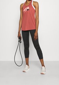 Punainen Nikellä varustettu toppi, valkoisten olkaimien päällä, yhdistetty mustiin capri-legginsseihin ja vaaleisiin lenkkareihin. Henkilöllä on käsissään hyppynaru.