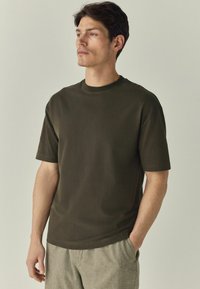 Olijfgroene t-shirt met korte mouwen en ronde hals, gemaakt van gladde stof. De pasvorm is ontspannen, met enkele minimale stikseldetails.