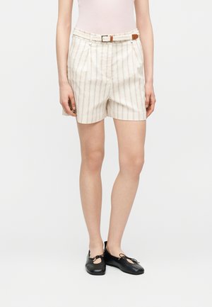 Nő, aki bézs színű rövidnadrágot visel vékony függőleges csíkokkal, fonott övet, fekete balettcipőt és világos, ujjatlan felsőt.