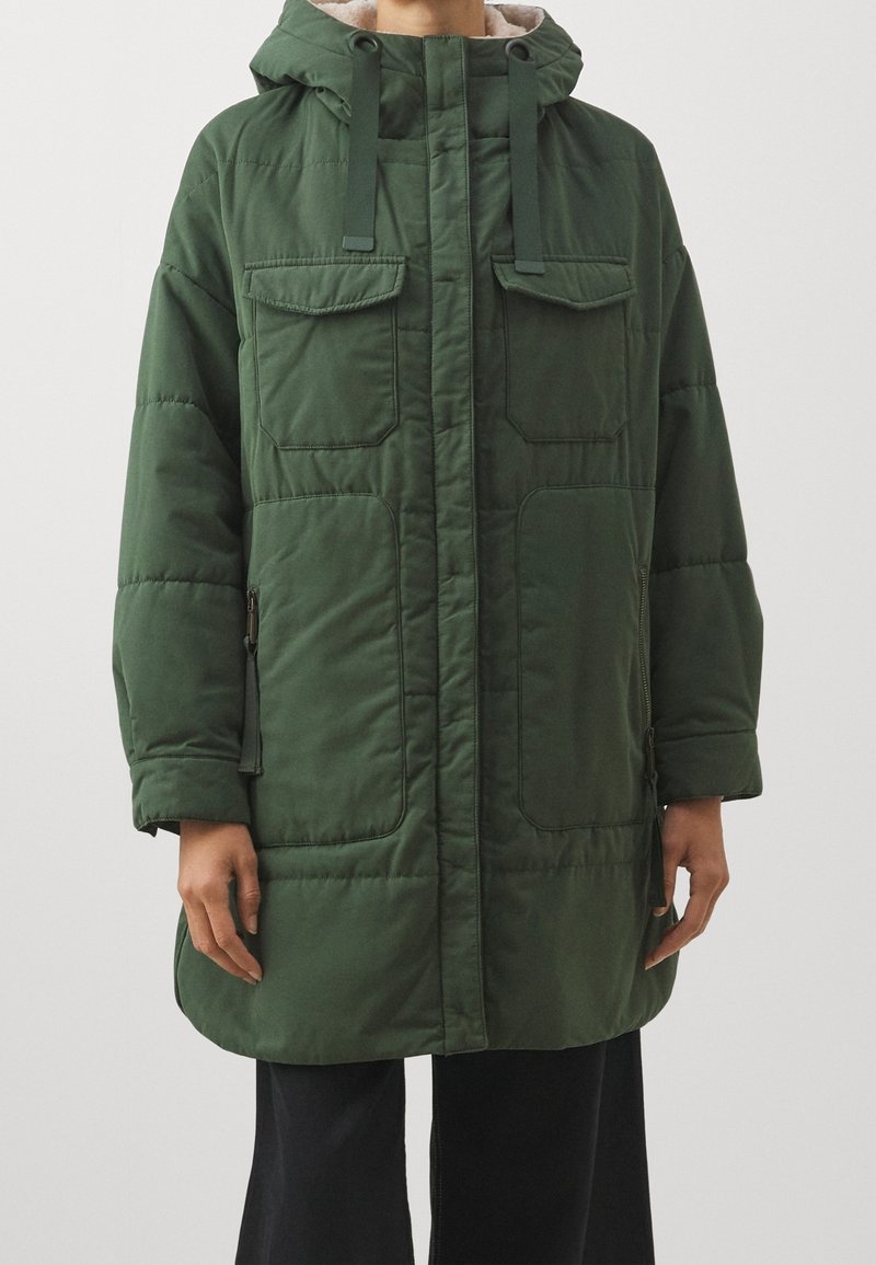 Grön vadderad jacka med huva, som har två bröstfickor, sidofickor med dragkedja och ett mjukt innerfoder. Tyget har en slät yta.