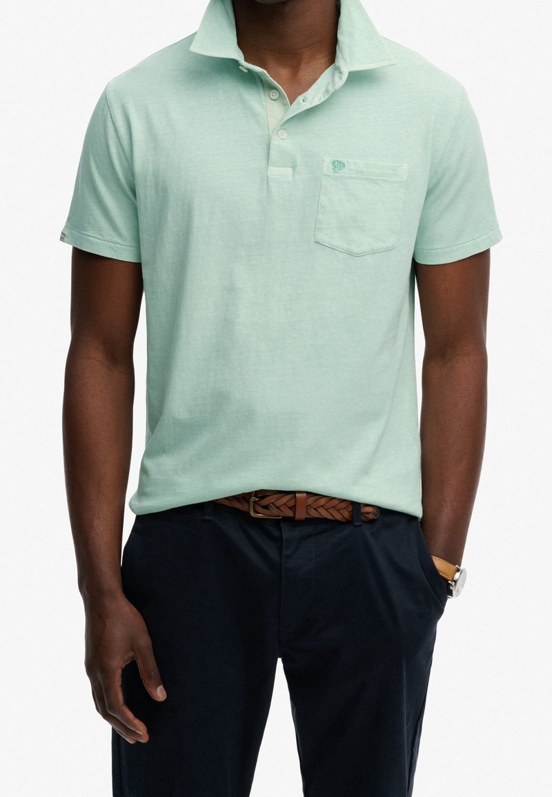 Homme portant un polo vert clair à manches courtes avec poche poitrine, pantalon sombre, ceinture marron tressée et montre au poignet gauche.