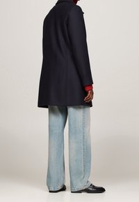 Marineblauer Wollmantel mit geradem Schnitt, hohem Kragen und langen Ärmeln; kombiniert mit hellblauen weiten Jeans und schwarzen Slippern.