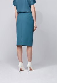 En figursydd turkos penskjol med en bakslits, tillsammans med vita högklackade ankelskor. Tyget har en slät, något reflekterande yta.