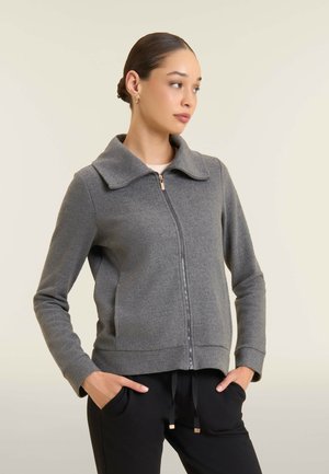 Giacca grigia con zip e collo grande, realizzata in tessuto morbido. Presenta tasche laterali e polsini a costine, abbinata a pantaloni neri.