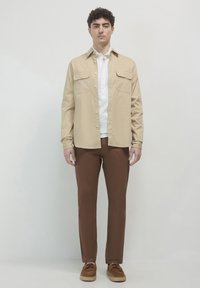 Helle beige Langarmshirt mit Knöpfen und Brusttaschen, über einem weißen Polo getragen. Kombiniert mit braunen Hosen und braunen Schuhen. Einfaches Design.