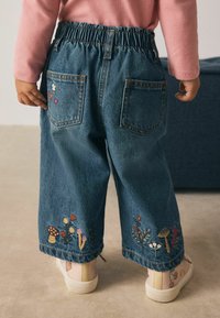 Kind, das blaue Jeans mit floralem und pilzförmigem Stickmuster auf den Beinen und einem rosa Langarmoberteil trägt, steht auf einem beigen Boden.