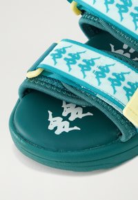 Tealfärgade sandaler med ett texturerat rem med ett upprepat mönster, en vadderad sula och en vit logotyp tryckt på sulan.