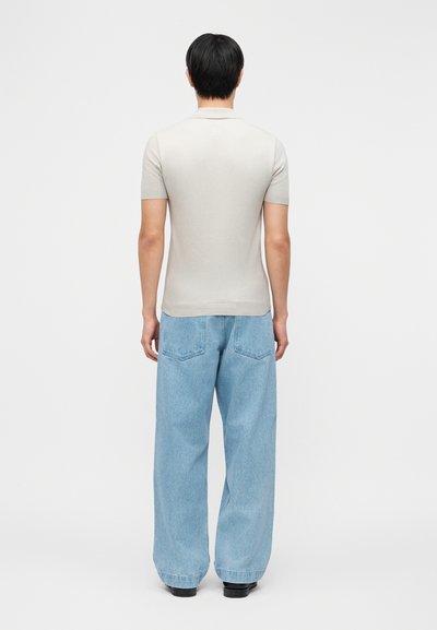 Άνδρας που στέκεται με την πλάτη προς τα εμπρός, φοράει μπεζ κοντομάνικο πόλο πουκάμισο και ανοιχτό μπλε τζιν παντελόνι με φαρδιά μπατζάκια και μαύρα παπούτσια σε απλό φόντο.