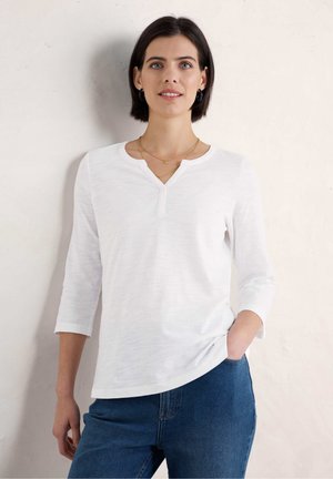 Witte, langemouwen top met een henley-halslijn, zachte textuur, lichte glans, gecombineerd met blauwe denim jeans, staande tegen een lichte achtergrond.