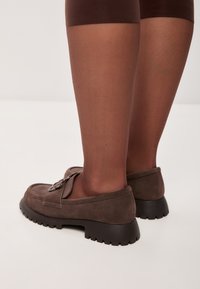 Mocassini in suede marrone con suola in gomma spessa e dettaglio a fiocco decorativo; indossati con collant sheer marroni, che mostrano una texture liscia.