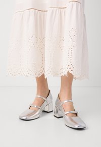 Zapatos Mary Jane metálicos plateados con punta cuadrada, dos correas y tacón cuadrado, combinados con un vestido blanco con bordado y dobladillo en forma de ondas.
