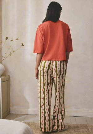 Žena stojaca vo vnútri s dlhými tmavými vlasmi, oblečená v voľnej oranžovej blúzke a vzorovaných nohaviciach so zelenými vlnitými pruhmi a hviezdičkovými motívmi.