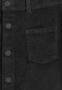 Schwarzer Cordstoff mit vertikaler Rippe, zwei runden Metallknöpfen und einer Brusttasche mit genähtem Rand.