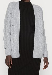 Grå stickad cardigan med texturerade, kabelstickade ärmar, öppen frontdesign, tillverkad av mjukt, fluffigt material, bärs över en svart randig klänning.